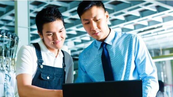 Two men standing together look at a laptop screen; one is dressed in business attire and the other is wearing overalls.