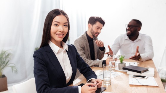 A woman in business attire sits at a conference table, smiling at the camera, while two men in the background talk and gesture.
