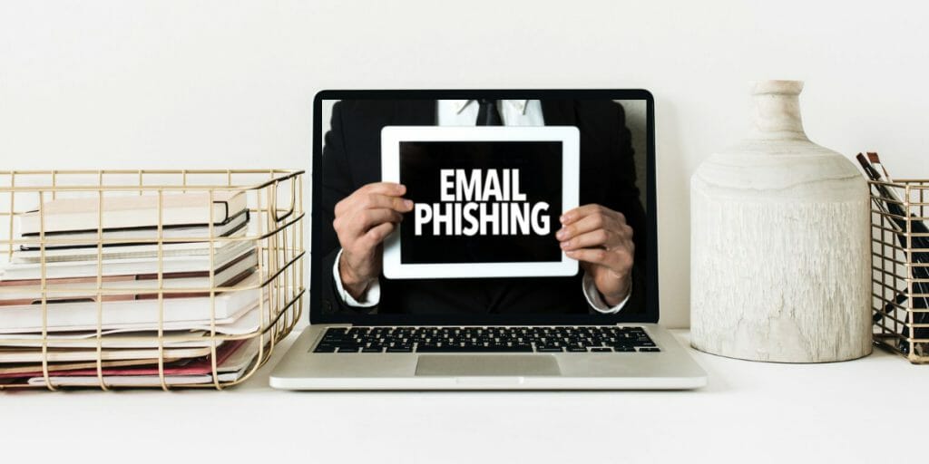 A laptop on a desk displays a person holding a tablet with the words "EMAIL PHISHING" on the screen, highlighting the need to protect my business against ransomware. Books and vases are placed on either side of the laptop.