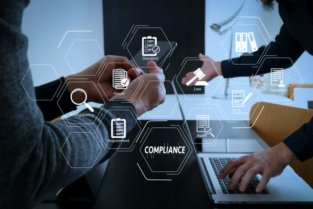 Two people working on a smartphone and laptop at a desk, with compliance-related icons digitally overlaid, including law, audit, IT Compliance, requirement, cybersecurity, and transparency.