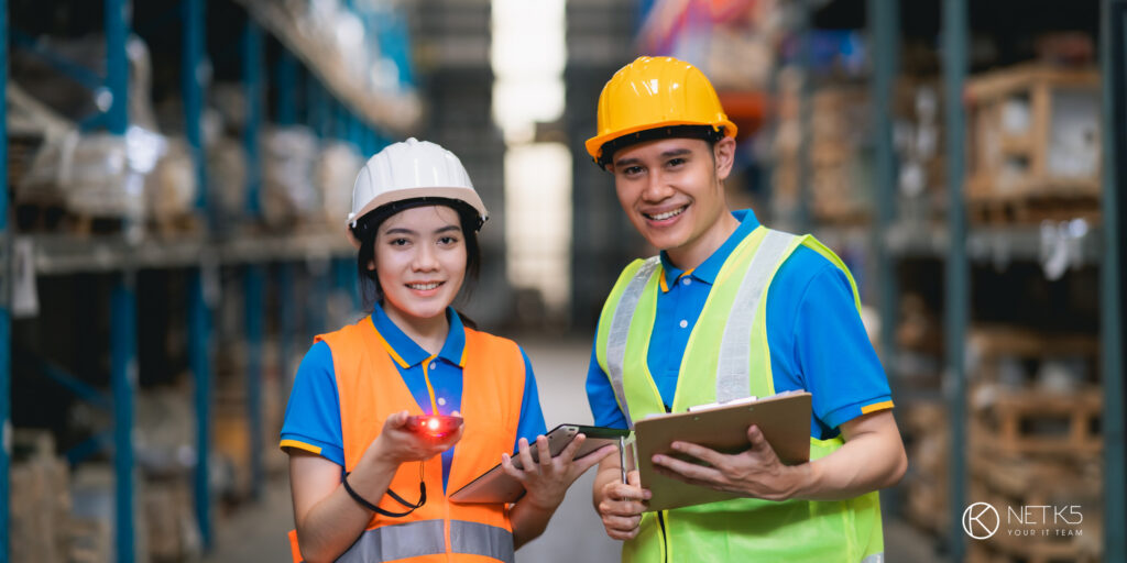Two warehouse workers in safety gear stand in an aisle, holding a clipboard and a barcode scanner, with shelves of inventory in the background.