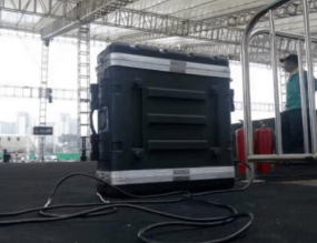 A large black equipment case with cables attached sits on a stage under a tent. Metal railings and a person in the background are visible.