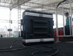 A large black equipment case with cables attached sits on a stage under a tent. Metal railings and a person in the background are visible.