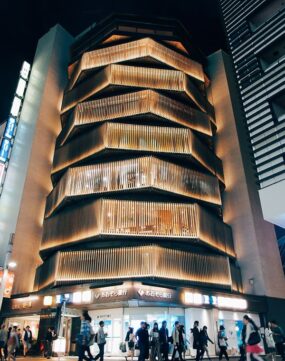 A modern, multi-story building with illuminated, layered wooden slats on the facade at night; people walk by on the street below.