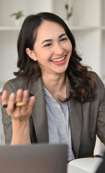 A woman in a blazer sits at a desk, smiling and gesturing with a pen in hand, with a notebook and laptop in front of her.