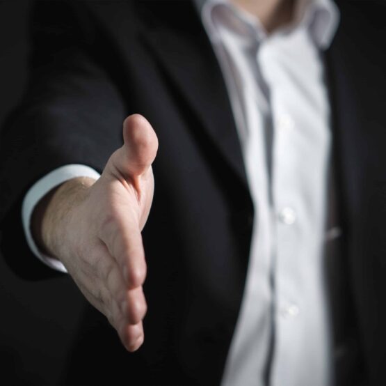 A person in a suit extends their hand forward, offering a handshake against a dark background. A person in a suit extends their hand forward, offering a handshake against a dark background.