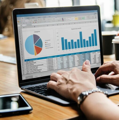 A person works on a laptop displaying spreadsheets with a pie chart and bar graph, while others sit around a table with papers, coffee cups, and a smartphone. A person works on a laptop displaying spreadsheets with a pie chart and bar graph, while others sit around a table with papers, coffee cups, and a smartphone.