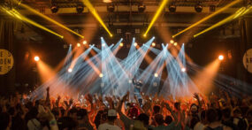 A large crowd with raised hands faces a stage with bright spotlights and a performer, in an indoor concert or music festival setting.